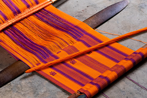 Image of orange and purple yarn being woven together on a wooden frame. The background is grey stone. Parts of the yarn are already woven and parts are in process.